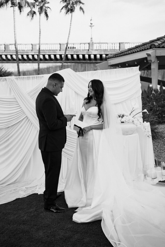 Bride reading handwritten vows to groom in front of a white draped ceremony backdrop during a modern, minimal wedding in Arizona.