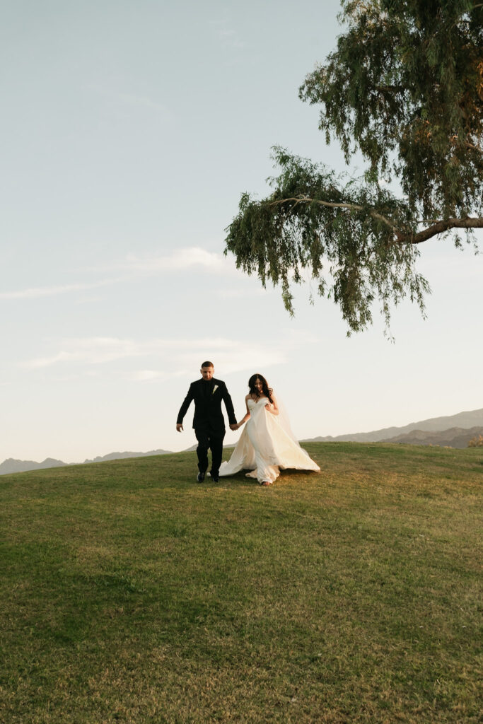 Bride and groom running together up a grassy hill beneath a large tree at sunset during their Arizona wedding photos.