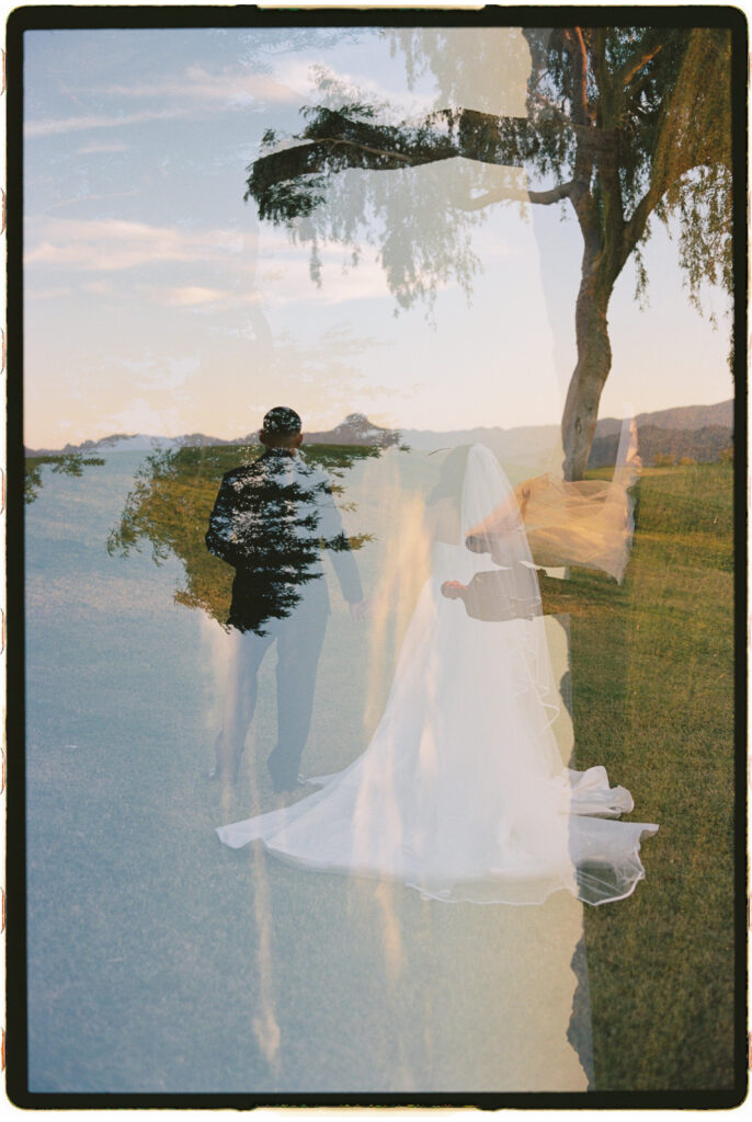 Bride and groom walking together up a grassy hill beneath a large tree at sunset during their Arizona wedding photos.